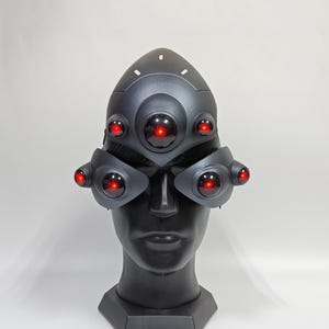 May include: A black helmet with red glowing lights. The helmet has a rounded top and a visor with multiple red lights. The helmet is on a black mannequin head.