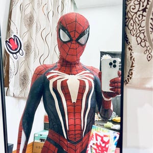 May include: A red and black Spider-Man costume with a white spider emblem on the chest. The costume features a web pattern and white eye lenses. The person is holding a white smartphone.