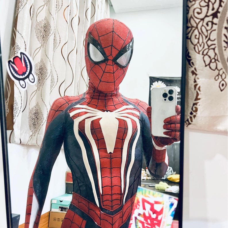 May include: A red and black Spider-Man costume with a white spider emblem on the chest. The costume features a web pattern and white eye lenses. The person is holding a white smartphone.