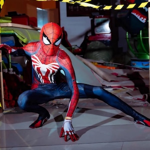 May include: A person in a Spider-Man costume, crouching low with one leg extended. The costume is red and blue with a white spider emblem on the chest. The mask has white eye lenses. The background is a cluttered indoor space.