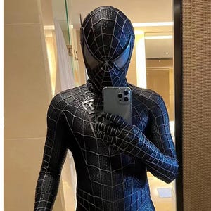 May include: A person wearing a black and blue spandex suit with a black spider web design. The person is holding a silver smartphone in front of a mirror.