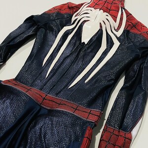 May include: A Spider-Man costume featuring a navy blue and red color scheme. The suit has a textured pattern and a large white spider emblem on the back. The costume includes a red belt and red and black checkered sections.