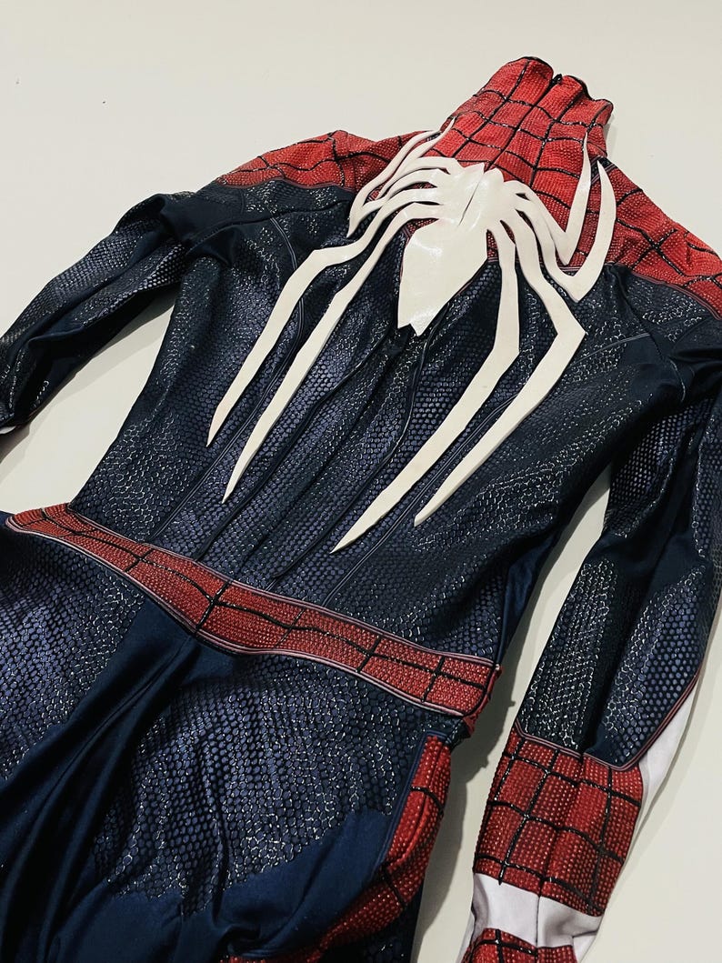 May include: A Spider-Man costume featuring a navy blue and red color scheme. The suit has a textured pattern and a large white spider emblem on the back. The costume includes a red belt and red and black checkered sections.