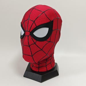 May include: A red Spider-Man mask with black webbing and large white eye lenses. The mask is displayed on a black stand. The mask is a replica of the superhero's mask.