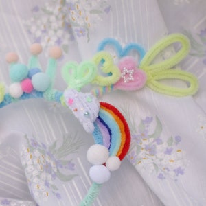 May include: A colorful headband with a rainbow, a flower, and a cloud design. The headband is made of pipe cleaners and pom-poms.