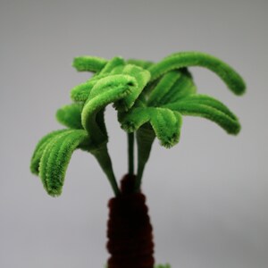 May include: A small green artificial palm tree in a white geometric pot. The palm tree is made of fuzzy green pipe cleaners and the pot has a textured surface.