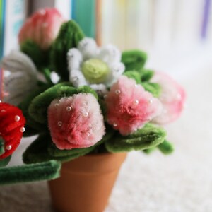 May include: A small potted plant with pink and white flowers made from pipe cleaners. The flowers are decorated with white beads.