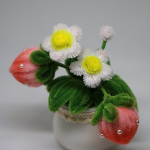 May include: A small white ceramic pot with a lace trim, filled with a faux flower arrangement. The arrangement features pink and white felt flowers with green felt leaves.