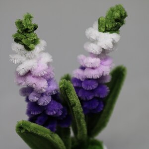 May include: Two handmade pipe cleaner flowers with green stems and leaves. The flower heads transition from purple to lavender, white, and green. The flowers are set against a soft gray background.