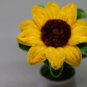 May include: A yellow felt sunflower with a brown felt center. The sunflower is in a small white pot with green felt leaves.