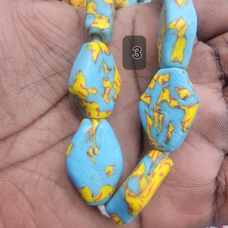 May include: Close-up of a strand of colorful beads. The beads are a light blue color with yellow and red patterns. The beads are strung together on a white string. The number "3" is visible in a white box.