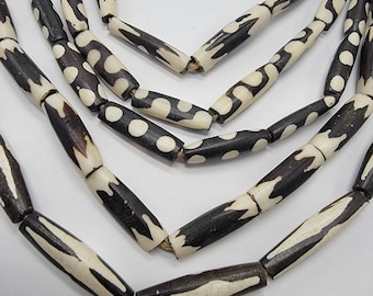 May include: A necklace made of black and white bone beads. The beads are cylindrical and have a geometric pattern. The necklace is strung on a gold cord.