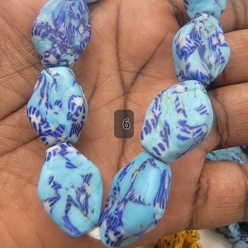 May include: A strand of large, irregular beads. The beads are a light blue color with a pattern of dark blue and white swirls. The beads are held in a hand.