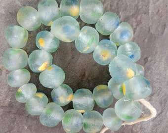 May include: A strand of translucent, light blue glass beads. Each bead has a slightly irregular shape and contains patches of yellow, green, and blue. The beads are strung on a white cord, creating a unique jewelry component.