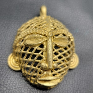May include: A gold-colored pendant featuring a detailed face design with a latticework pattern. The face has a prominent nose and closed eyes, with a textured surface.