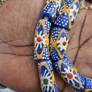 May include: Close-up of a strand of colorful glass beads. The beads are primarily blue with white polka dots and yellow and white floral designs. Each bead has a central red or green dot.