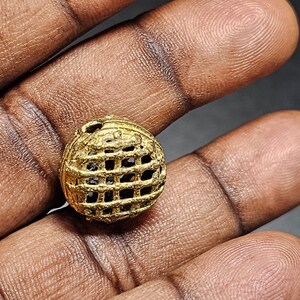 May include: A gold-colored metal bead with a geometric, grid-like design. The bead is round and has a hollow center.