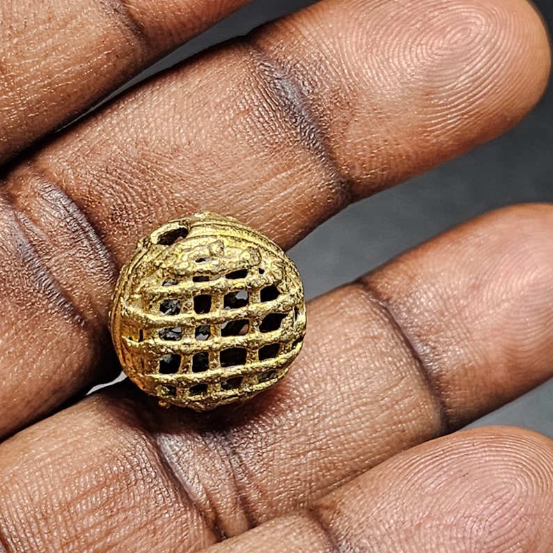 May include: A gold-colored metal bead with a geometric, grid-like design. The bead is round and has a hollow center.