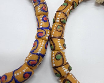 May include: Two beaded bracelets with intricate patterns. The beads are a light brown color with white, blue, green, and red painted designs. The bracelets are joined together at the center.