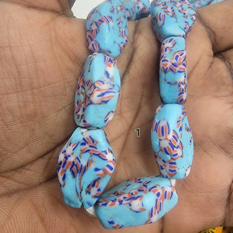 May include: A strand of light blue, irregularly shaped beads with a pattern of red, white, and blue stripes. The beads are strung together on a white cord. The number "1" is visible on the image.