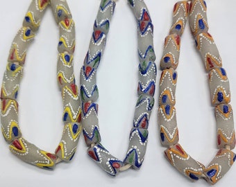 May include: Three strands of gray glass beads with colorful geometric patterns. The beads are strung on a white cord and have a white, blue, red, and yellow paint design.
