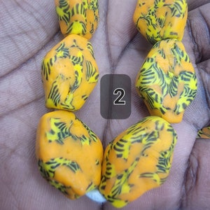 May include: Close-up of a strand of vibrant orange beads with a unique pattern of black and yellow stripes. The beads have an irregular, organic shape. The number "2" is in a black square.