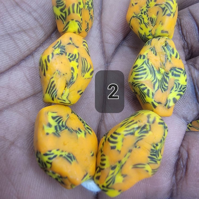 May include: Close-up of a strand of vibrant orange beads with a unique pattern of black and yellow stripes. The beads have an irregular, organic shape. The number "2" is in a black square.