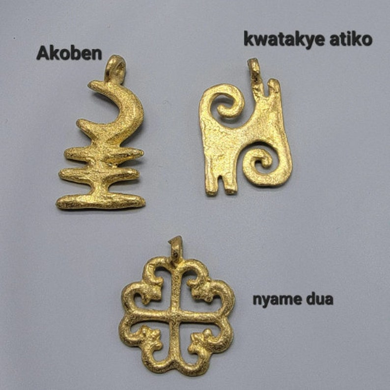 May include: Three gold-colored pendants with different designs. The pendant on the left is labeled "Akoben", the one in the middle is labeled "kwatakye atiko", and the one on the right is labeled "nyame dua".