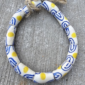 May include: A white ceramic necklace with blue and yellow painted designs. The necklace is made of cylindrical beads with a repeating pattern of blue swirls and yellow dots.