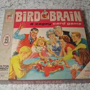 May include: A vintage board game box with a red, white, and blue color scheme. The box features a colorful illustration of a family playing the game, with the title "Bird Brain: a cagey card game" printed in large, bold letters. The game is for 2 to 4 players and is recommended for ages 8 to adult.