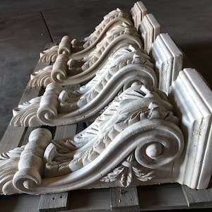 May include: Four ornate, white concrete corbels with intricate scrollwork and leaf designs. The corbels are stacked on top of each other, showcasing their detailed craftsmanship.
