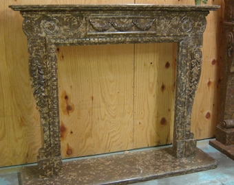 May include: An antique-style fireplace mantel in shades of brown and tan. The mantel features ornate carvings and a rectangular base. A small plant sits on the top of the mantel. The fireplace is made of a stone-like material.