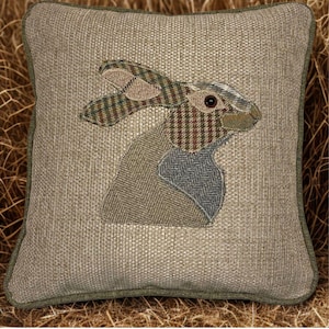 Puede incluir: Cojín decorativo cuadrado con diseño de conejo. El cojín presenta una tela texturizada de tonos neutros con un borde verde. El conejo está hecho de varias telas estampadas en tonos marrones, verdes y grises.