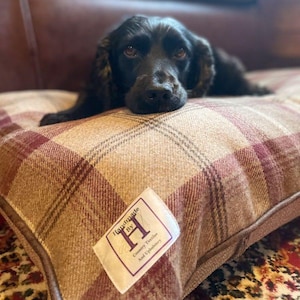 May include: A dog bed with a plaid pattern in shades of beige and burgundy. The bed has a rectangular shape and a label that reads "Handmade By H Country Textiles And Upholstery". A black dog is resting on the bed.
