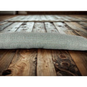 May include: A light grey, cylindrical door draft excluder made of a textured fabric. The excluder is lying on a wooden floor with a natural wood grain pattern. The excluder is designed to block draughts.