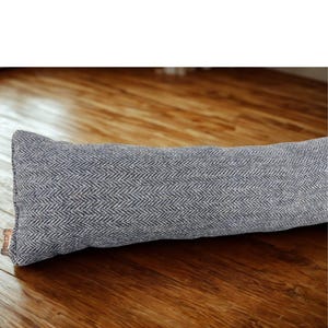 May include: A gray and white herringbone pattern door draft stopper. The rectangular pillow-shaped stopper is made of a textured fabric. The stopper is resting on a wooden floor.