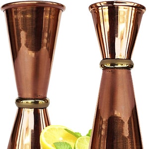 Könnte beinhalten: Zwei kupferfarbene Jigger mit einem goldenen Band, zum Messen von Flüssigkeiten. Eine Zitrone, eine Limette und Minzzweige befinden sich im Vordergrund. Die Jigger haben einen ausgestellten oberen Teil und eine konische Basis.