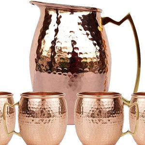 May include: A copper pitcher and four matching copper mugs with gold-colored handles. The pitcher has a hammered texture, and the mugs have a similar hammered finish. The set is designed for serving beverages.