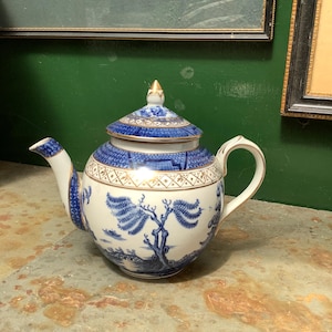 May include: A blue and white porcelain teapot with a lid, spout, and handle. The teapot has a blue and gold geometric pattern around the rim and lid, and a blue tree and landscape design on the body. The teapot is on a stone surface.