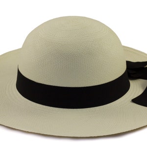 Pu&ograve; includere: Un cappello Panama beige chiaro con un'ampia tesa e un nastro nero attorno alla corona. Il nastro ha un fiocco su un lato.