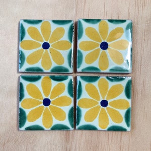 May include: Four square ceramic tiles with a floral design. Each tile features a yellow flower with a blue centre on a white background, framed by a green border. The tiles are arranged on a light brown wooden surface.