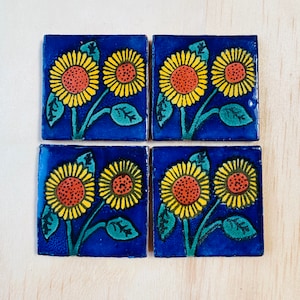 Op de afbeelding: Vier vierkante keramische tegels met een zonnebloemontwerp. Elke tegel heeft een diepblauwe achtergrond met twee zonnebloemen in geel en oranje, groene bladeren en stelen.