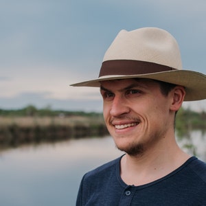 Könnte beinhalten: Ein Mann mit einem hellbraunen Fedora mit braunem Band, der lächelnd in die Kamera schaut. Er steht vor einem Gewässer mit grüner Vegetation im Hintergrund.