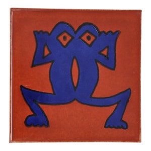 Op de afbeelding: Een blauwe gestileerde kikker met twee ogen op een rode achtergrond. De kikker is zwart omlijnd.