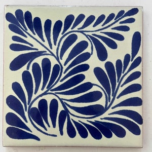 May include: A white ceramic tile with a blue floral pattern. The pattern features stylized leaves with rounded edges, creating a symmetrical design.