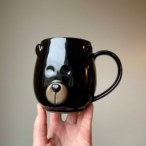 Peut inclure: Mug en céramique noire avec un visage d'ours. La tasse a une finition brillante et un nez brun.