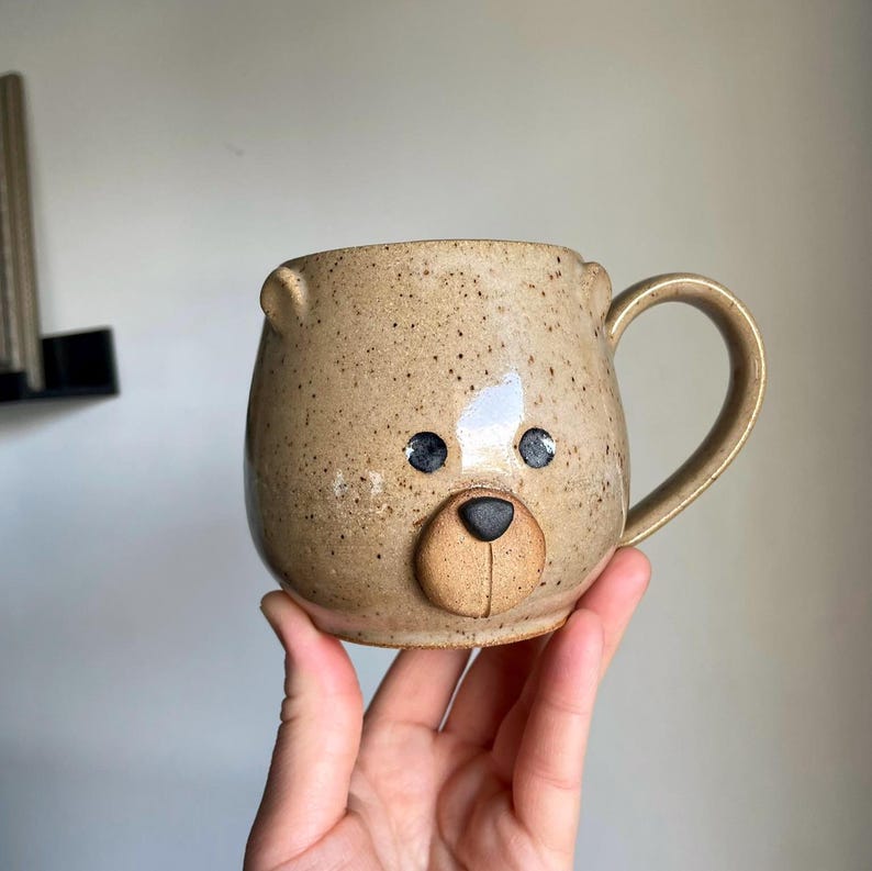 Puede incluir: Una taza de cer&aacute;mica beige con forma de cara de oso, con un cuerpo redondeado, un asa y orejas peque&ntilde;as. La cara del oso presenta ojos negros y un hocico beige. La taza se sostiene en una mano.