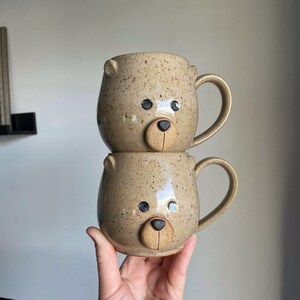 Puede incluir: Dos tazas de cer&aacute;mica hechas a mano apiladas con caras de oso. Las tazas son de color beige con motas oscuras, con orejas de oso, ojos negros y un hocico beige. Cada taza tiene un asa curvada.