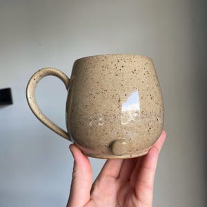 Puede incluir: Una taza de cer&aacute;mica beige moteada con un cuerpo redondeado y un asa grande. La taza tiene un peque&ntilde;o pie redondeado en la parte inferior. La taza est&aacute; sujeta por una mano.