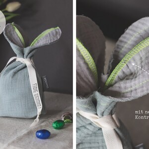 Puede incluir: Una bolsa de conejo de tela azul claro con una cinta blanca y una etiqueta que dice "Frohe Ostern!" La bolsa tiene costuras de color amarillo ne&oacute;n alrededor de las orejas.